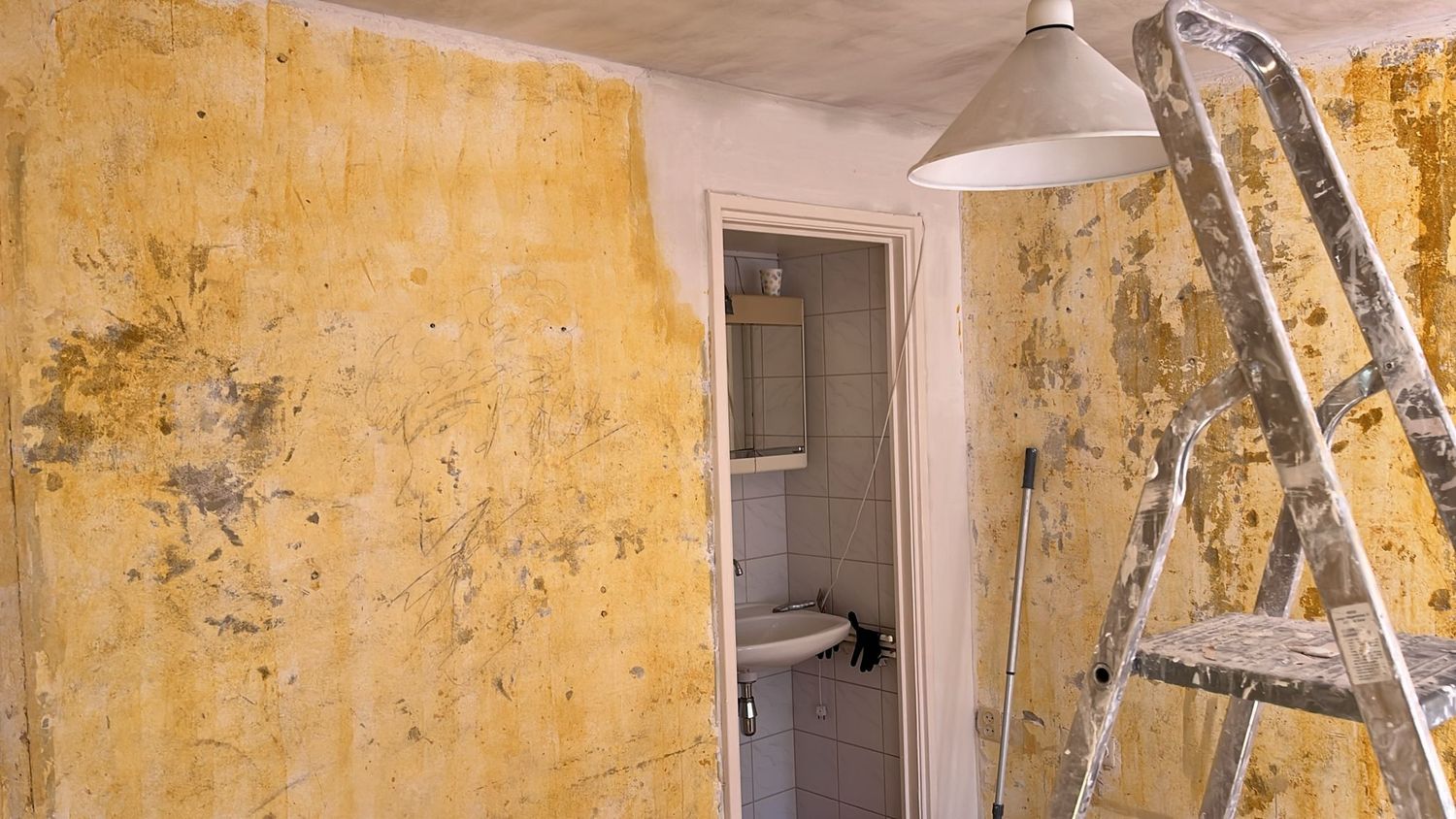 Halwand met okergele lijmresten naast een betegeld toilet